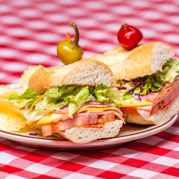 Two Italian subs with deli meats, cheese, lettuce, and peppers on a red and white checkered tablecloth.