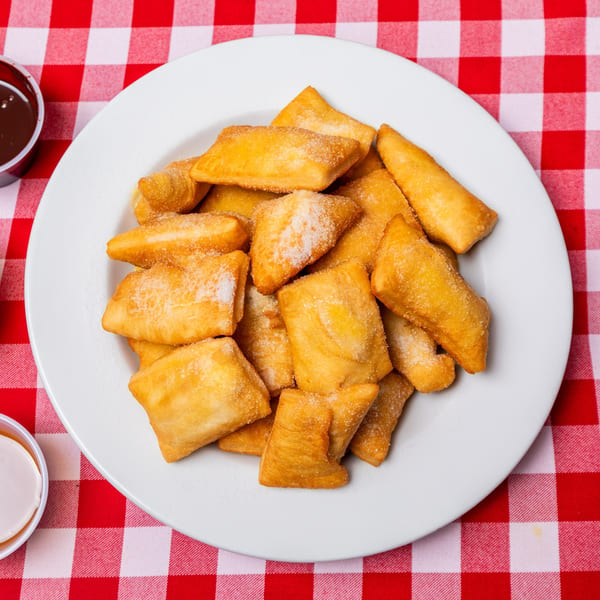 A white plate filled with golden-brown, sugar-dusted fried dough pieces on a red and white checkered tablecloth.